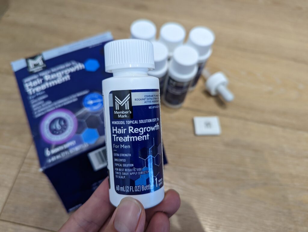 手持一罐 Member's Mark 5% Minoxidil 生髮水，特寫展示深藍色與白色相間的正面標籤，標示 60ml 單月容量。