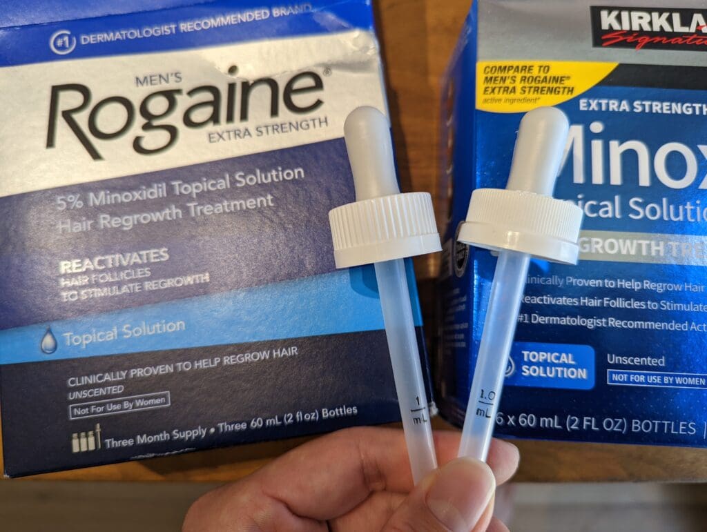 Rogaine 落建與 Kirkland 生髮水原廠滴管實體對比特寫：左側為 Rogaine，右側為 Kirkland。背景為各自的品牌外盒，展示 1mL 刻度線印刷清晰度與塑膠材質差異。 (